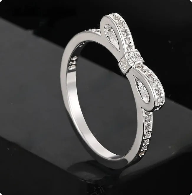 Silver coloured ring with a detailed bow shape set with clear sparkling zircon crystals on a model's hand.