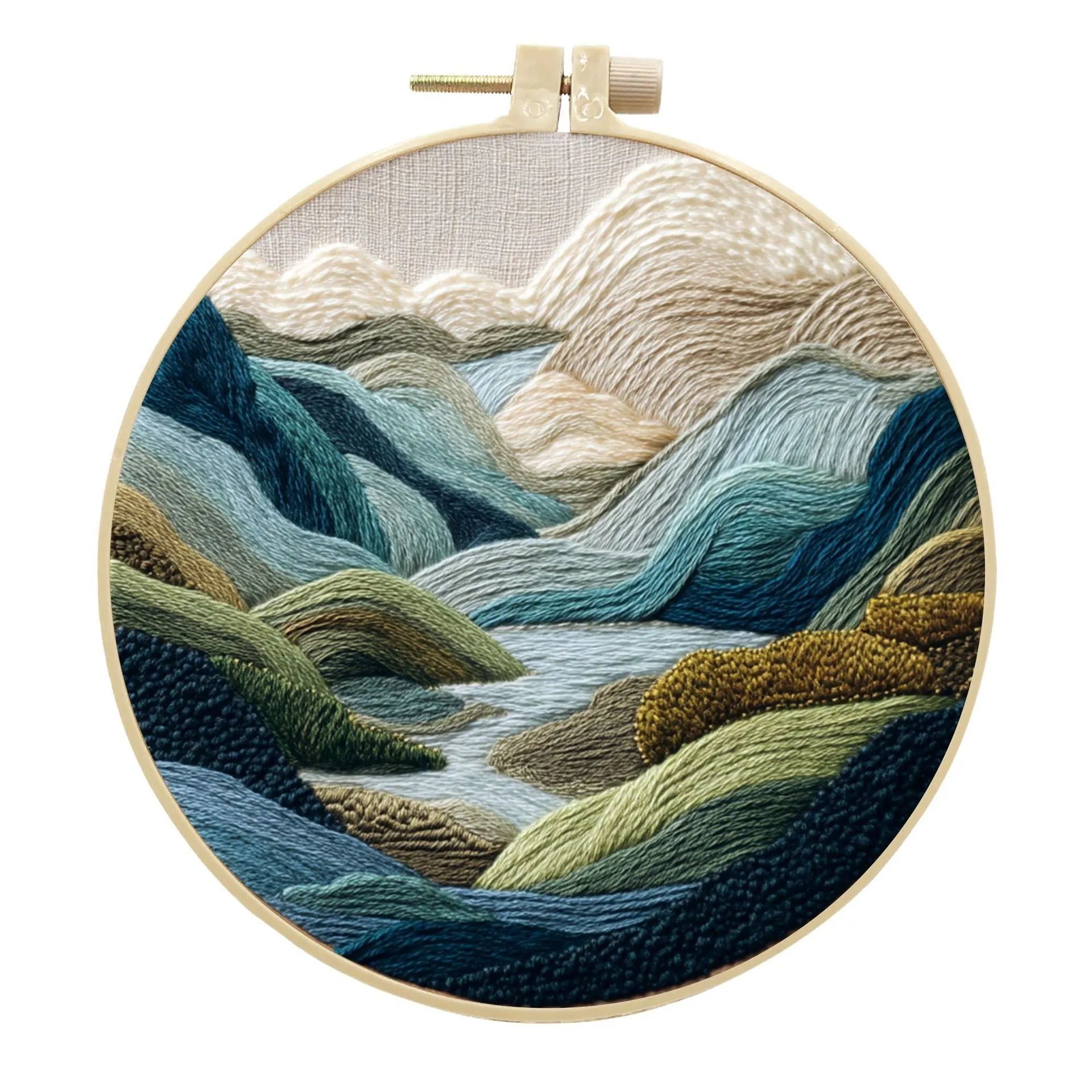Embroidery DIY kit components: printed landscape fabric, embroidery hoop, coloured threads, and needles laid flat.