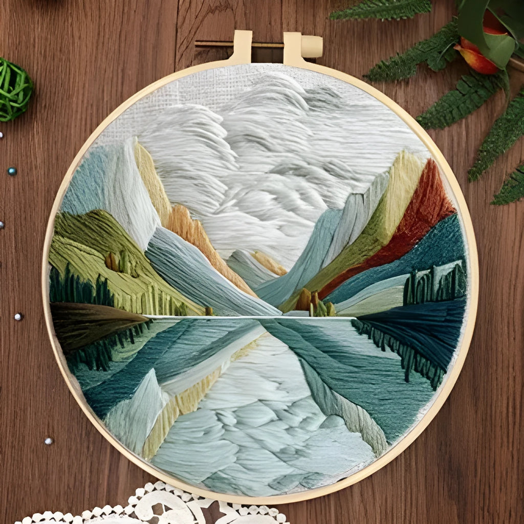 Embroidery DIY kit components: printed landscape fabric, embroidery hoop, coloured threads, and needles laid flat.