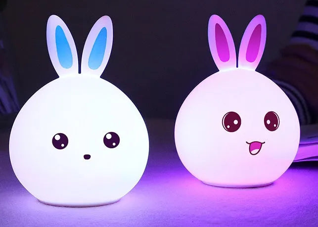 Cute rabbit-shaped night light made of soft silicone with touch sensor, showing seven LED colour options, on a bedside table.