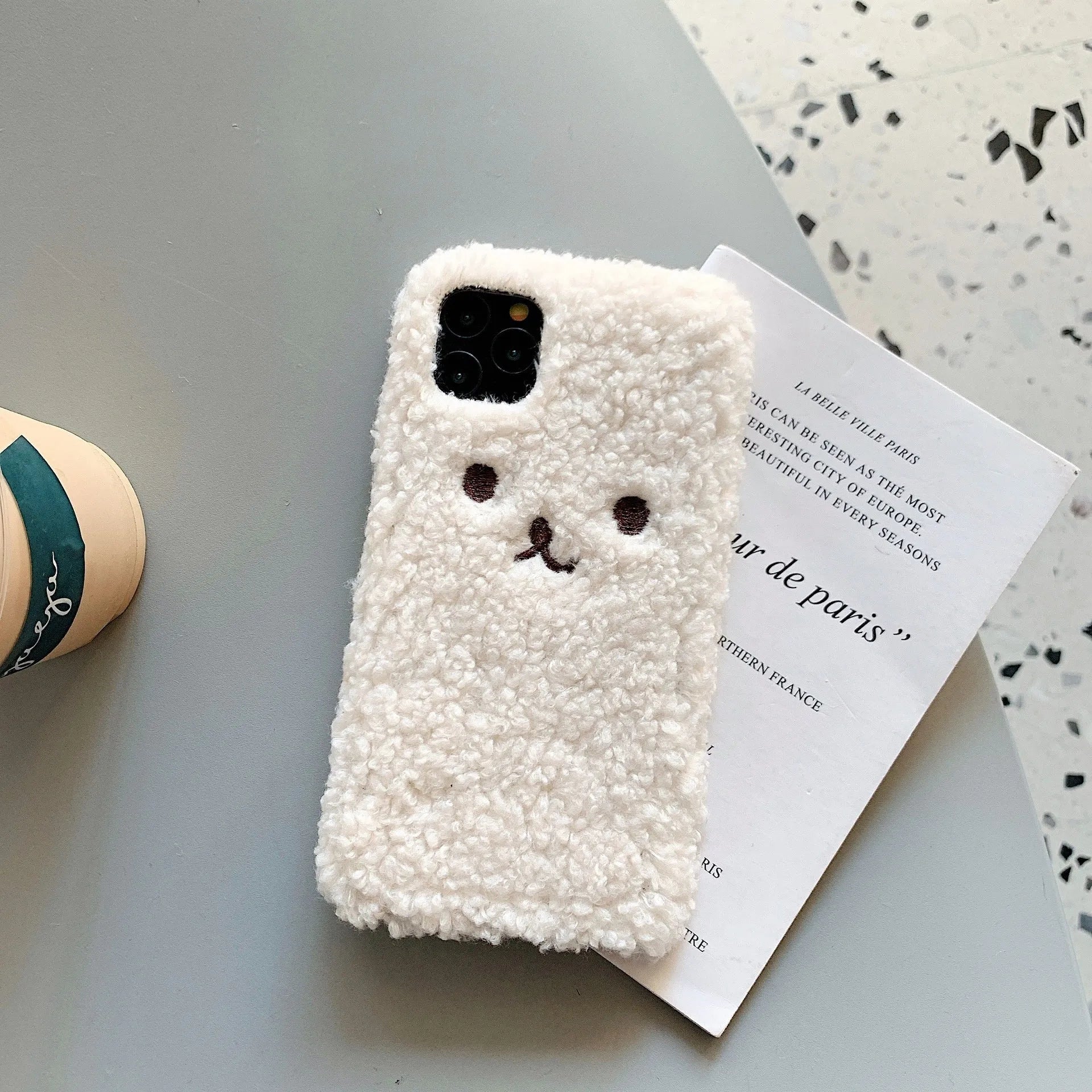 Brown embroidered plush bear face on a flexible TPU phone case for iPhone, showing camera cutout and raised edges.