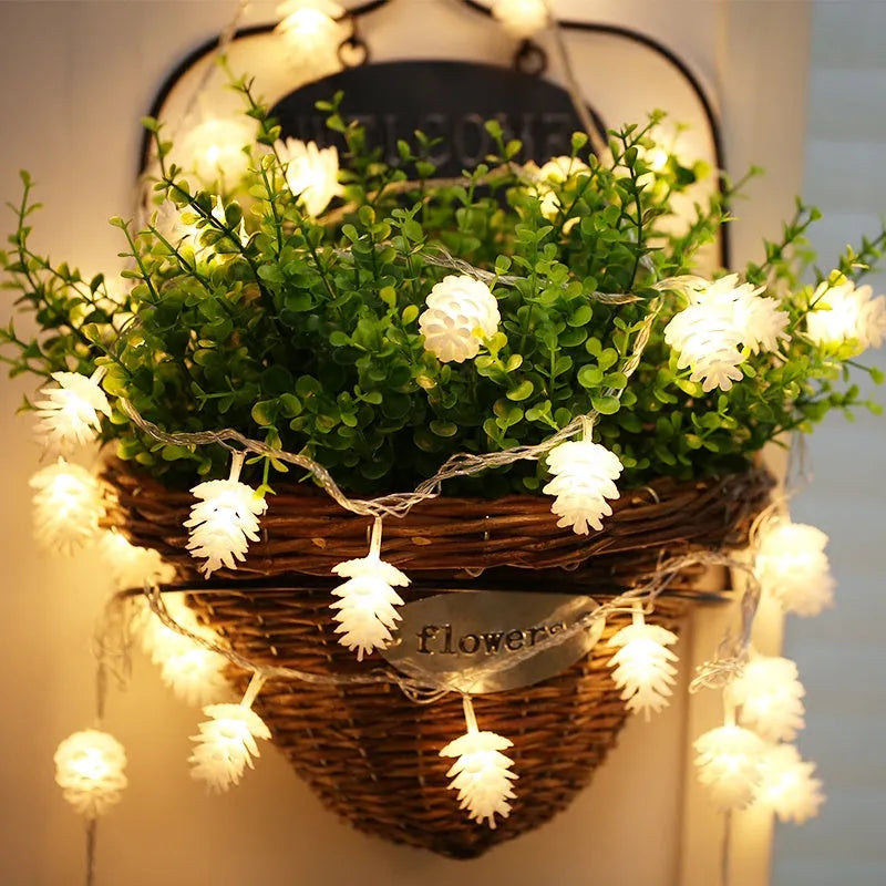Warm white LED pine cone decorative light string powered by batteries on a green foliage background.