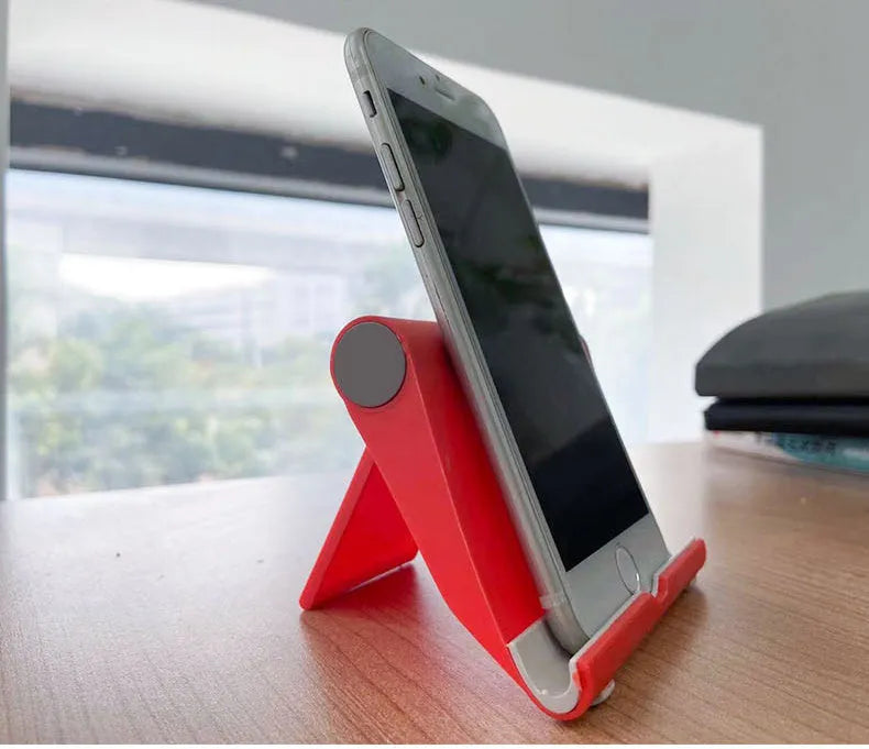 Black PE folding tablet stand holding a mobile phone at an adjustable viewing angle on a wooden desk.