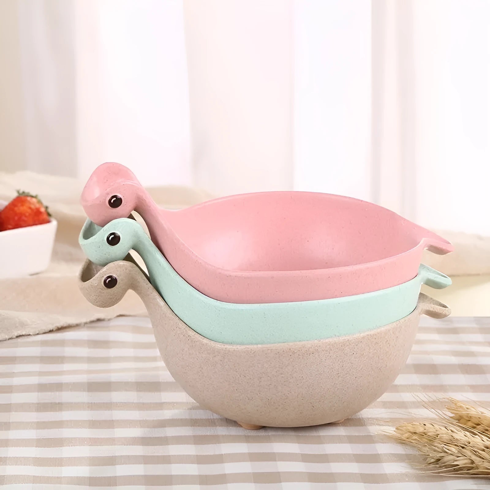 Three wheat straw dinosaur bowls in pink, green, and beige on a kitchen table