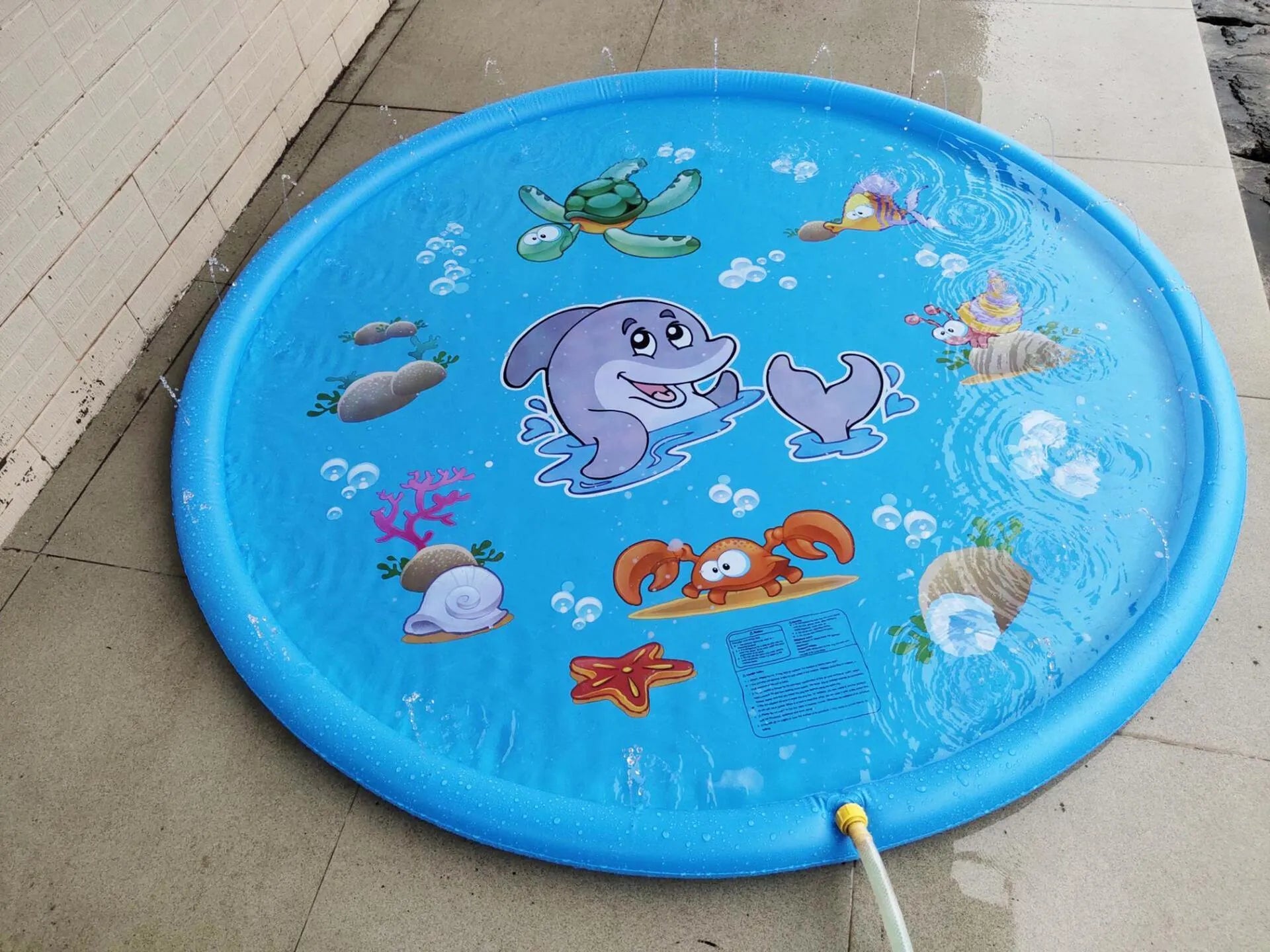 Blue children's water spray play mat with marine animal graphics on grass connected to a hose