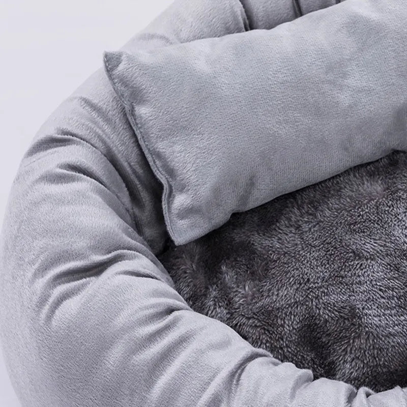 Grey round plush pet bed with raised edge and pillow, shown on a non-slip base.