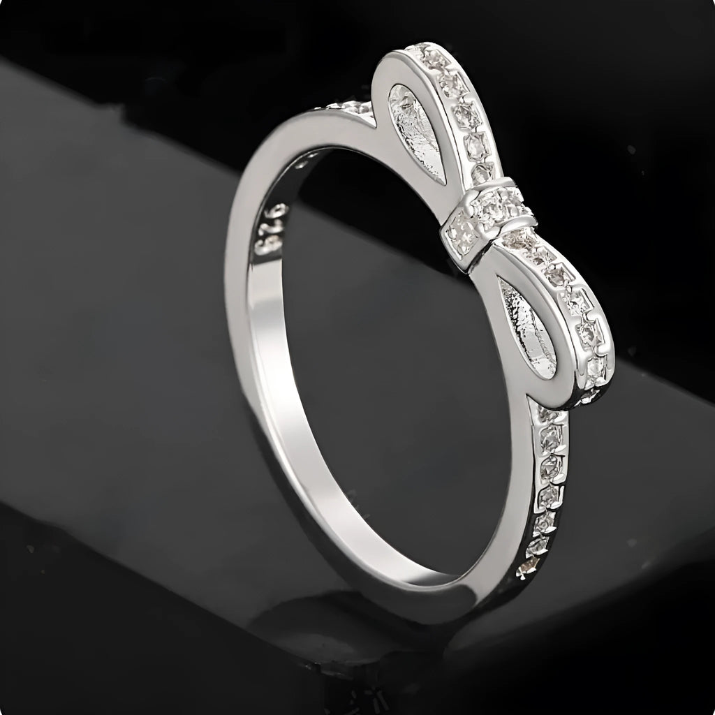 Silver coloured ring with a detailed bow shape set with clear sparkling zircon crystals on a model's hand.