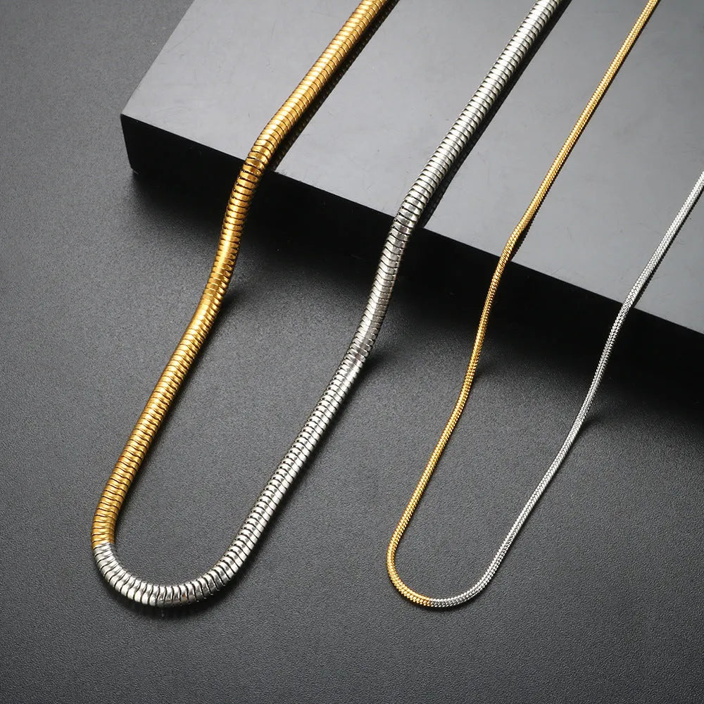 Close-up of a cold gold 3mm wide snake bone chain necklace with a geometric box clasp on a dark background.