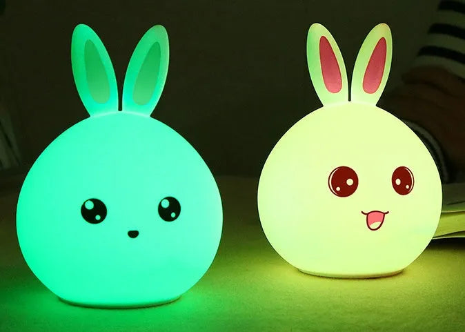 Cute rabbit-shaped night light made of soft silicone with touch sensor, showing seven LED colour options, on a bedside table.