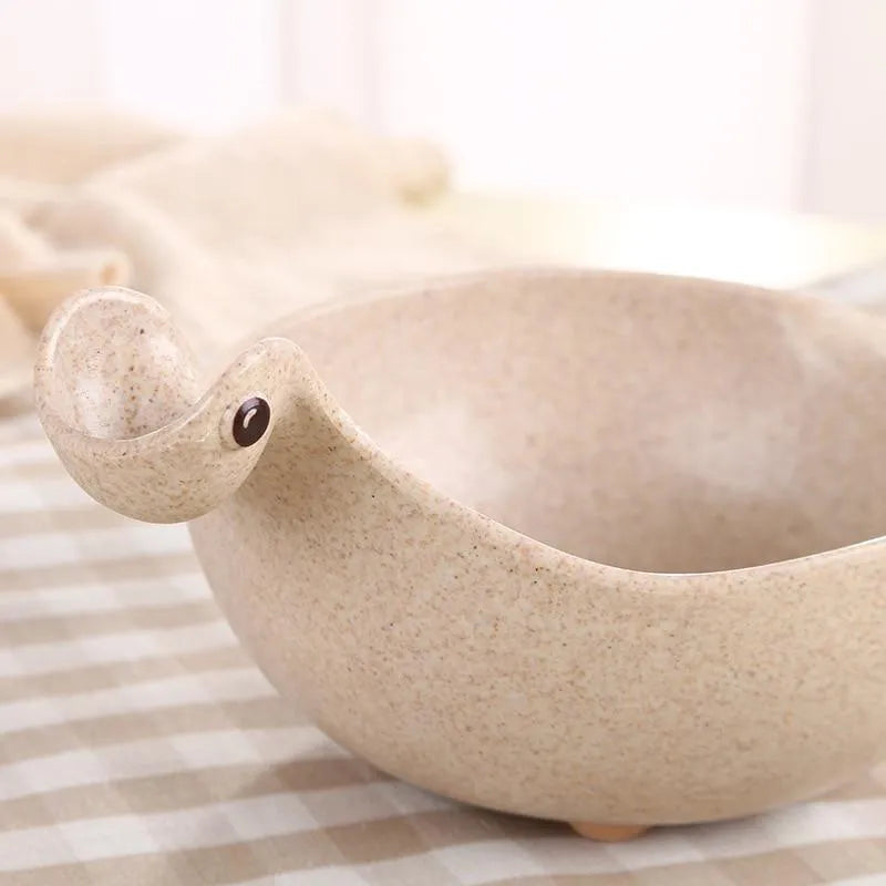 Three wheat straw dinosaur bowls in pink, green, and beige on a kitchen table