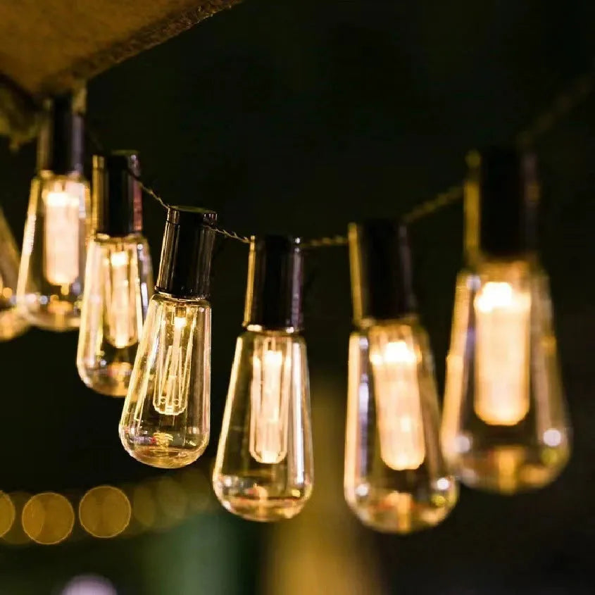 Outdoor retro long bulb infusion bottle shape string lights hanging in a garden setting.