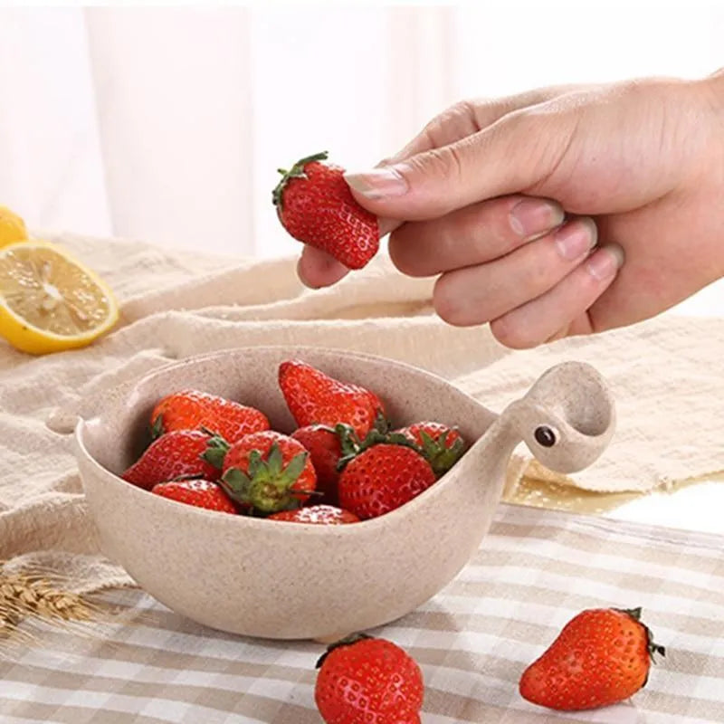 Three wheat straw dinosaur bowls in pink, green, and beige on a kitchen table