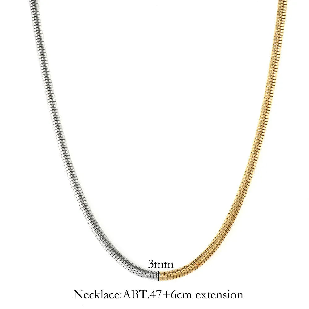 Close-up of a cold gold 3mm wide snake bone chain necklace with a geometric box clasp on a dark background.