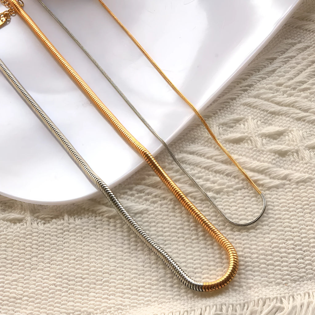 Close-up of a cold gold 3mm wide snake bone chain necklace with a geometric box clasp on a dark background.
