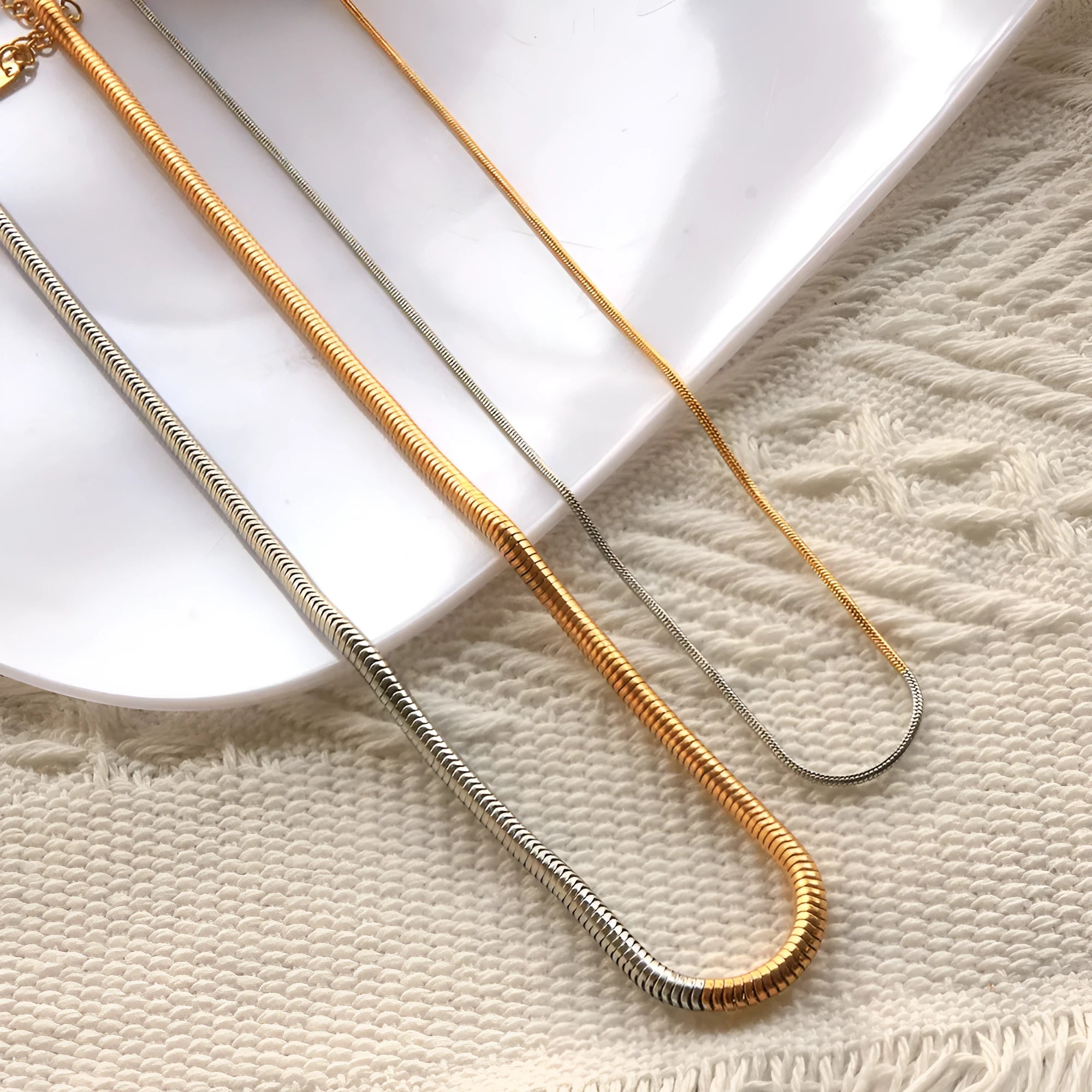 Close-up of a cold gold 3mm wide snake bone chain necklace with a geometric box clasp on a dark background.