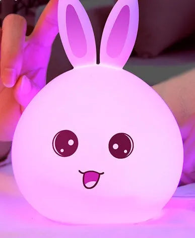 Cute rabbit-shaped night light made of soft silicone with touch sensor, showing seven LED colour options, on a bedside table.