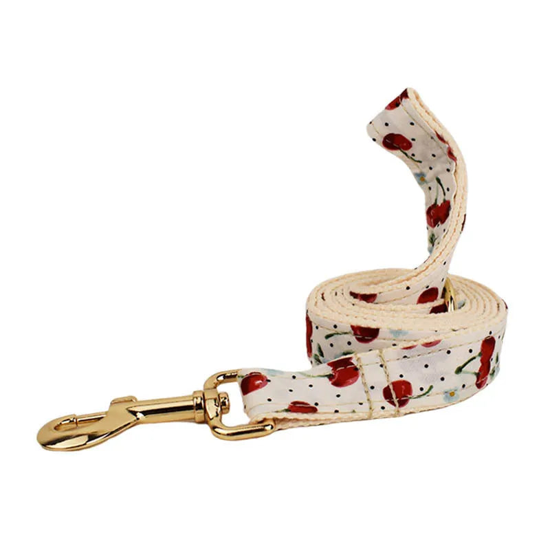 Red cherry print dog collar and leash set with light gold buckle, matching bow, vest, and poop bag.
