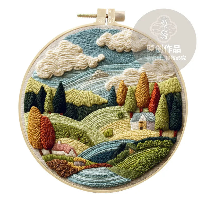 Embroidery DIY kit components: printed landscape fabric, embroidery hoop, coloured threads, and needles laid flat.