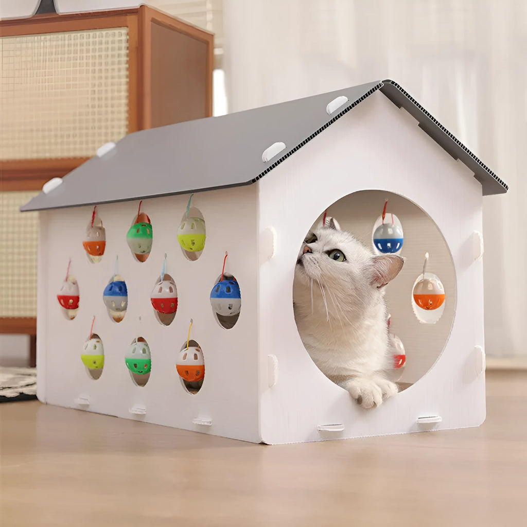 White plastic house-shaped cat scratch board with sisal siding, an arched tunnel entrance, and a dangling red bell ball toy.
