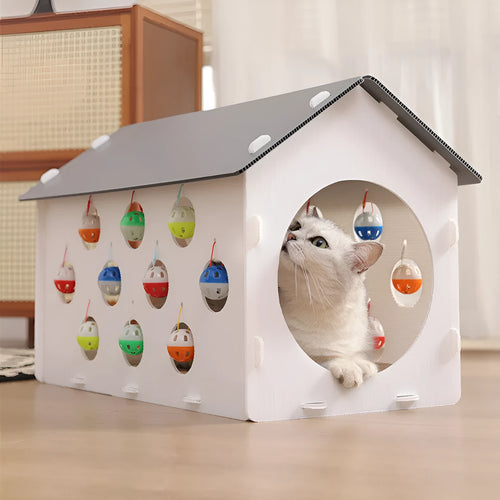 White plastic house-shaped cat scratch board with sisal siding, an arched tunnel entrance, and a dangling red bell ball toy.