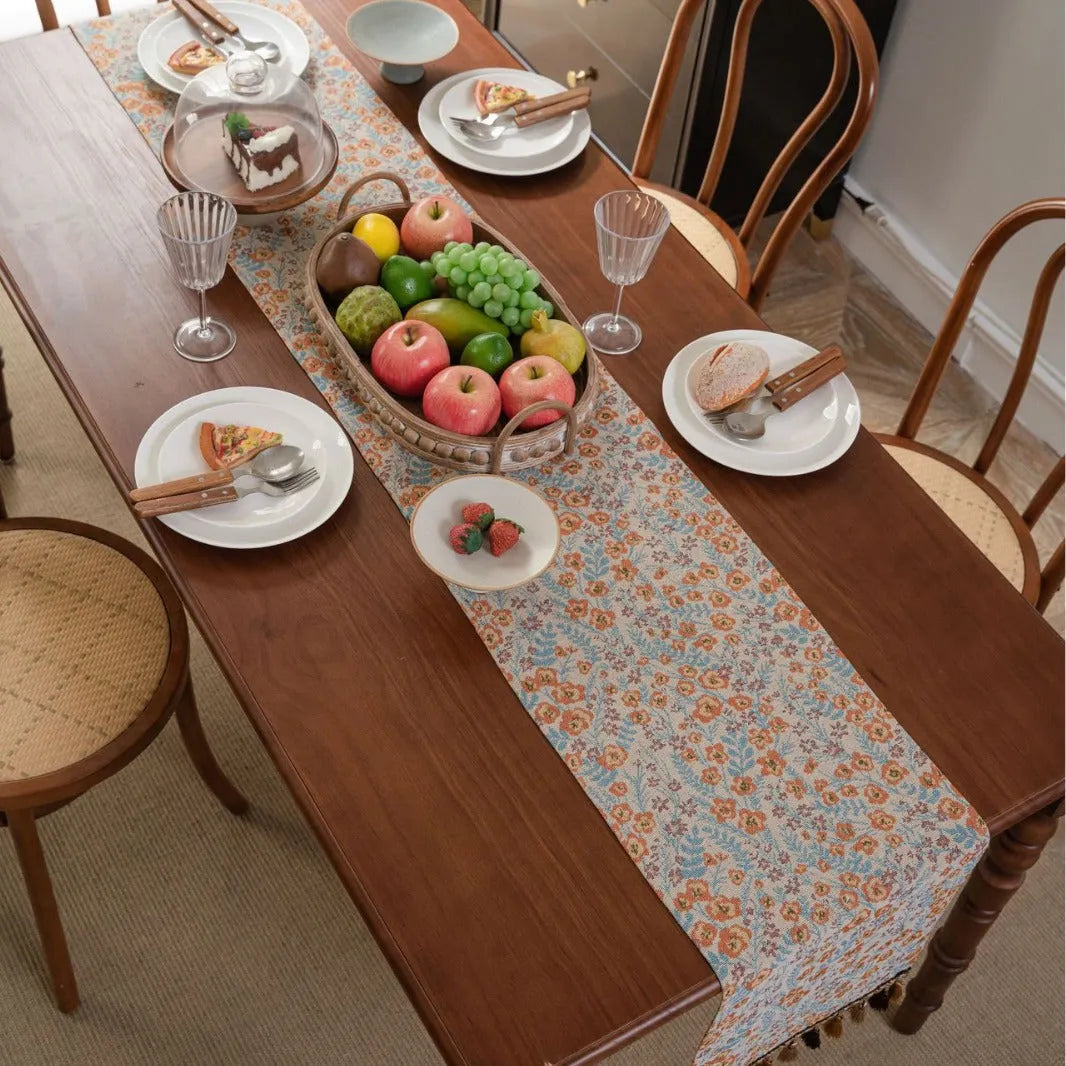 Beige jacquard table runner with floral pattern and long tassels on a wooden dining table.