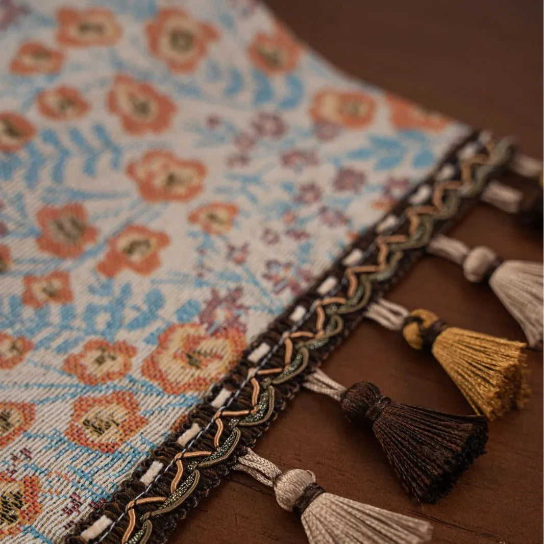 Beige jacquard table runner with floral pattern and long tassels on a wooden dining table.