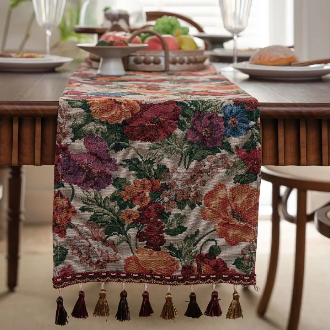 Beige jacquard table runner with floral pattern and long tassels on a wooden dining table.