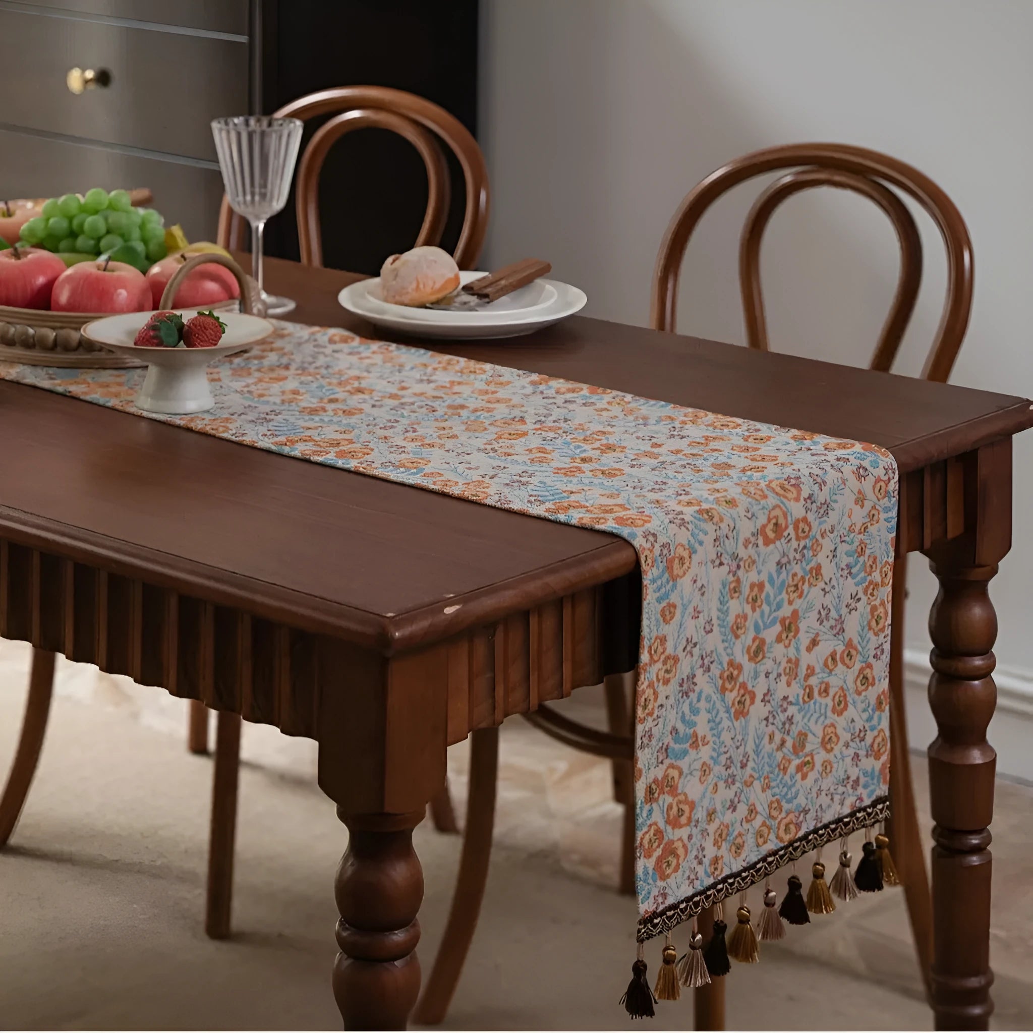 Beige jacquard table runner with floral pattern and long tassels on a wooden dining table.