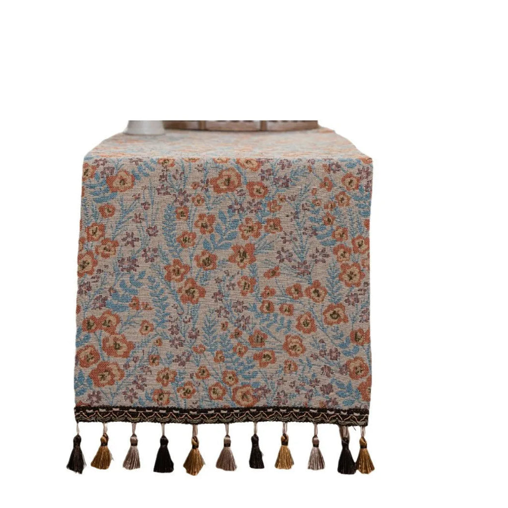 Beige jacquard table runner with floral pattern and long tassels on a wooden dining table.
