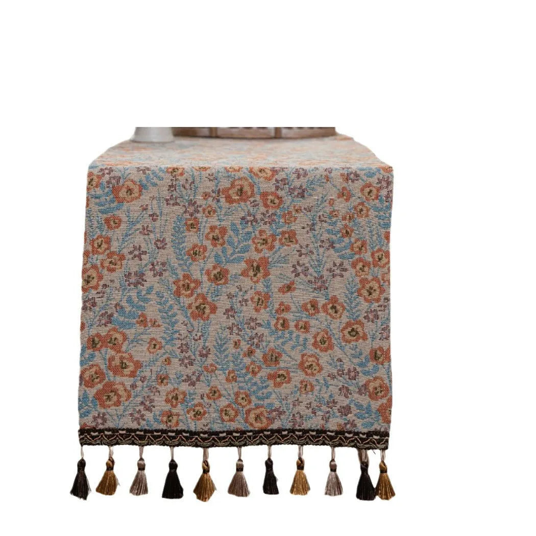 Beige jacquard table runner with floral pattern and long tassels on a wooden dining table.