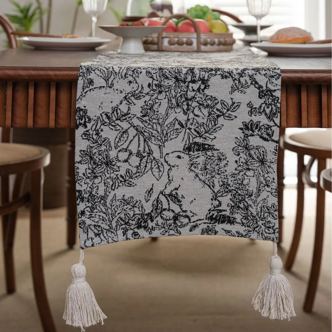 Beige jacquard table runner with floral pattern and long tassels on a wooden dining table.