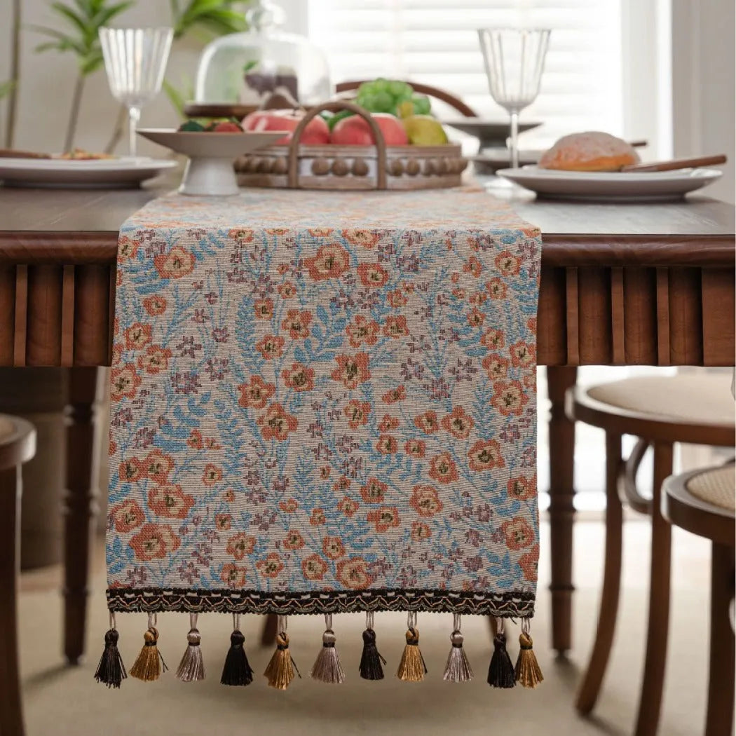 Beige jacquard table runner with floral pattern and long tassels on a wooden dining table.