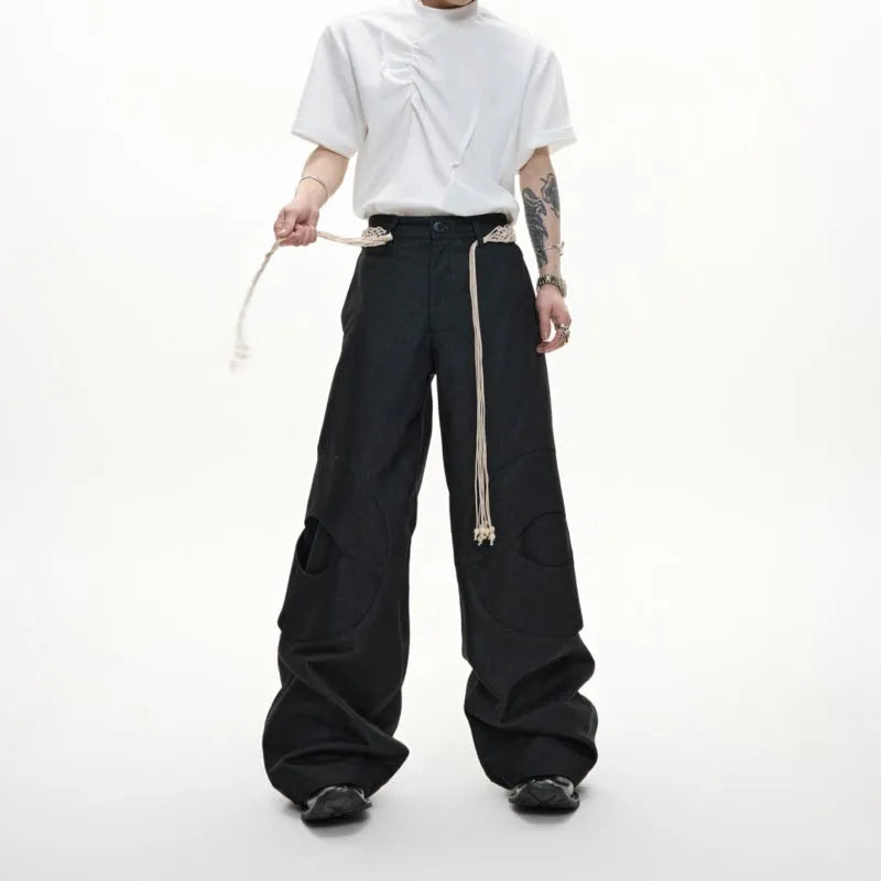 Man wearing dark blue cotton straight-leg trousers with heavy 3D belt stitching and decorative tassel details.