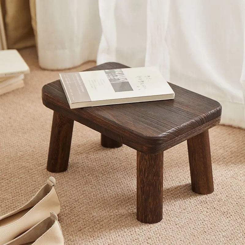 Distressed solid wood shoe changing stool with bracket legs in a living room setting.