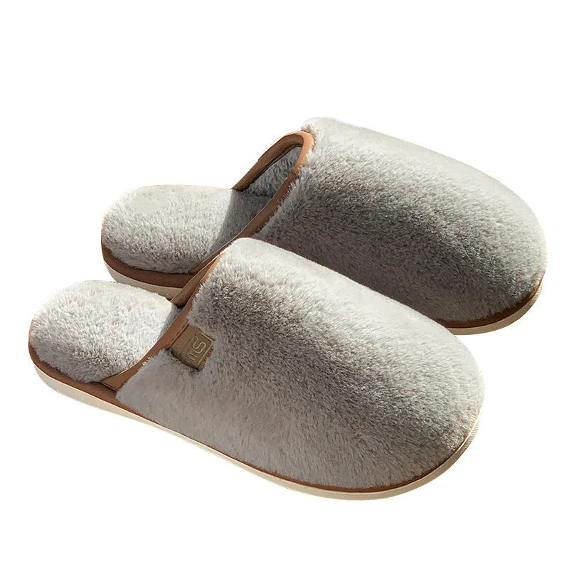 A pair of solid blue plush slip-on house slippers with a non-slip PVC sole and decorative car stitching on the upper.