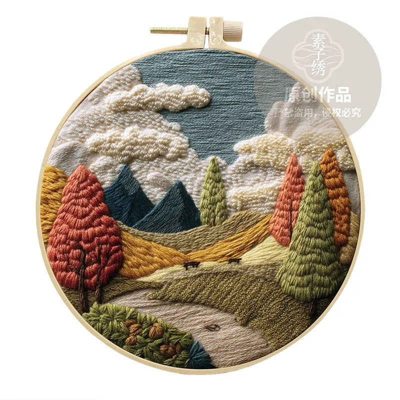 Embroidery DIY kit components: printed landscape fabric, embroidery hoop, coloured threads, and needles laid flat.
