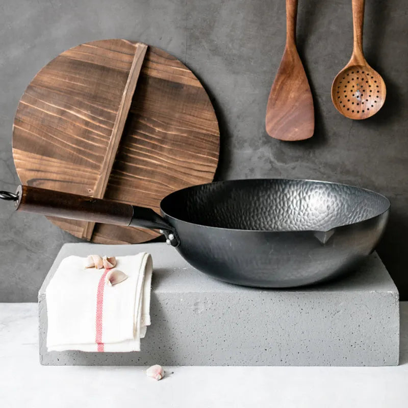 Traditional black cast iron wok with a long wooden handle, shown with a glass lid.