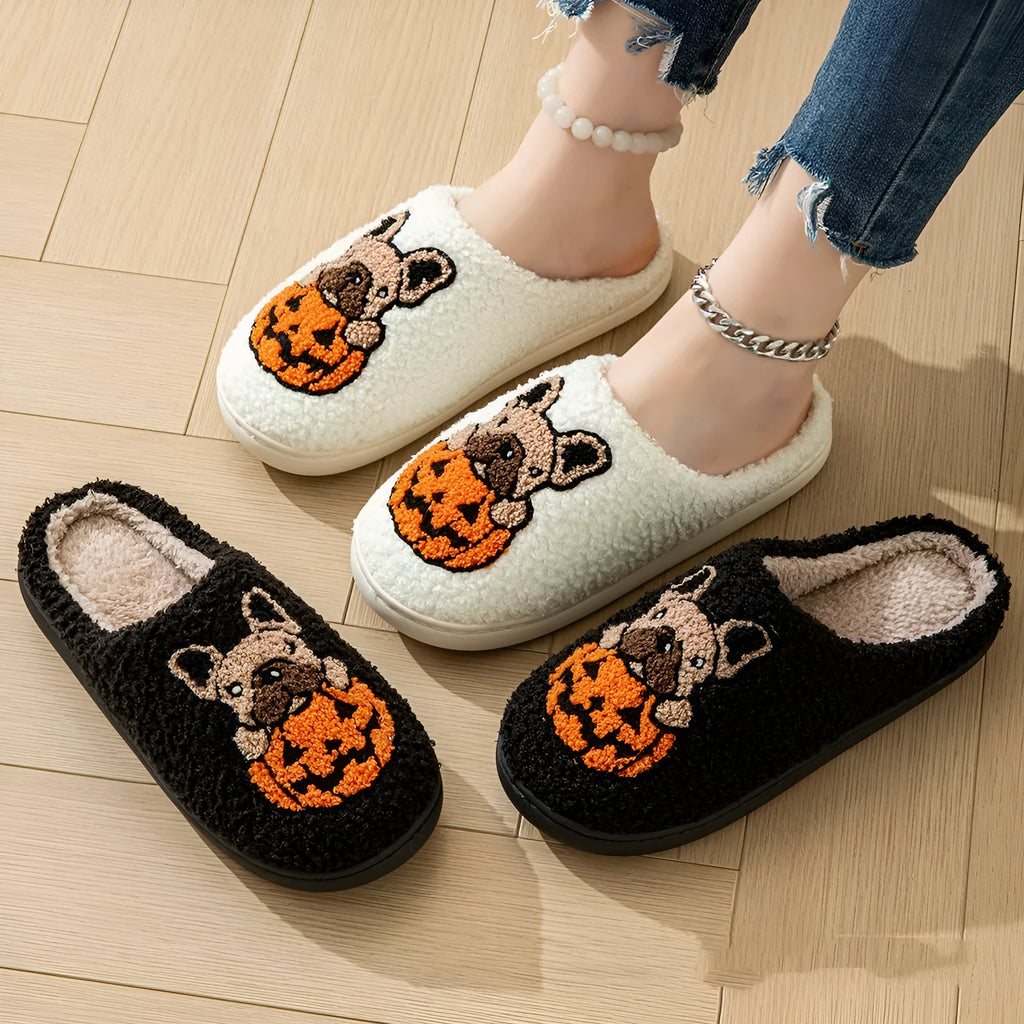 Black Halloween slippers with pumpkin print, plush fabric, and thick textured TPR rubber soles on a wooden floor.