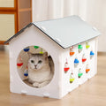 White plastic house-shaped cat scratch board with sisal siding, an arched tunnel entrance, and a dangling red bell ball toy.