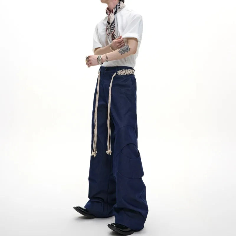 Man wearing dark blue cotton straight-leg trousers with heavy 3D belt stitching and decorative tassel details.