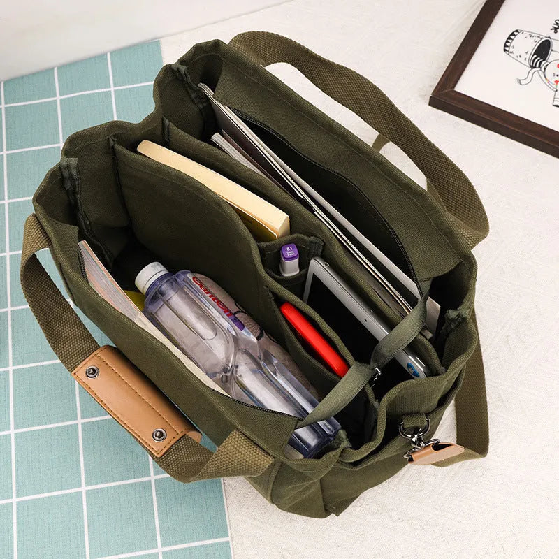 A horizontal square canvas tote bag in army green with a cross-body strap and visible sewing lines.