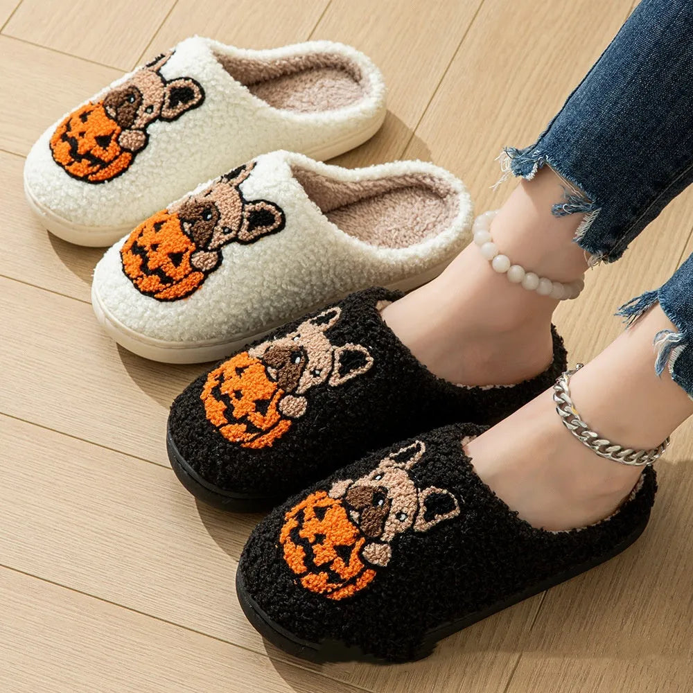 Black Halloween slippers with pumpkin print, plush fabric, and thick textured TPR rubber soles on a wooden floor.