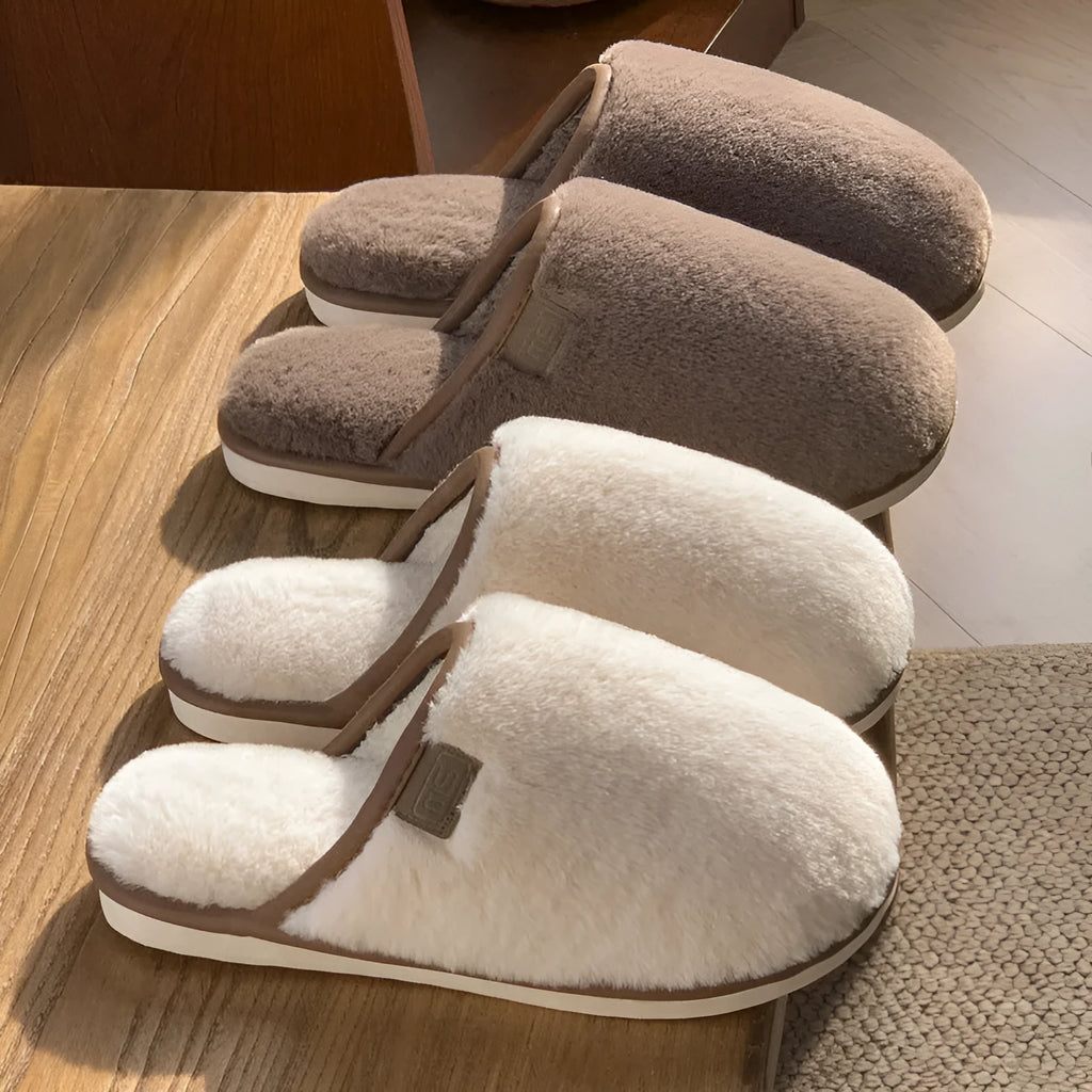 A pair of solid blue plush slip-on house slippers with a non-slip PVC sole and decorative car stitching on the upper.