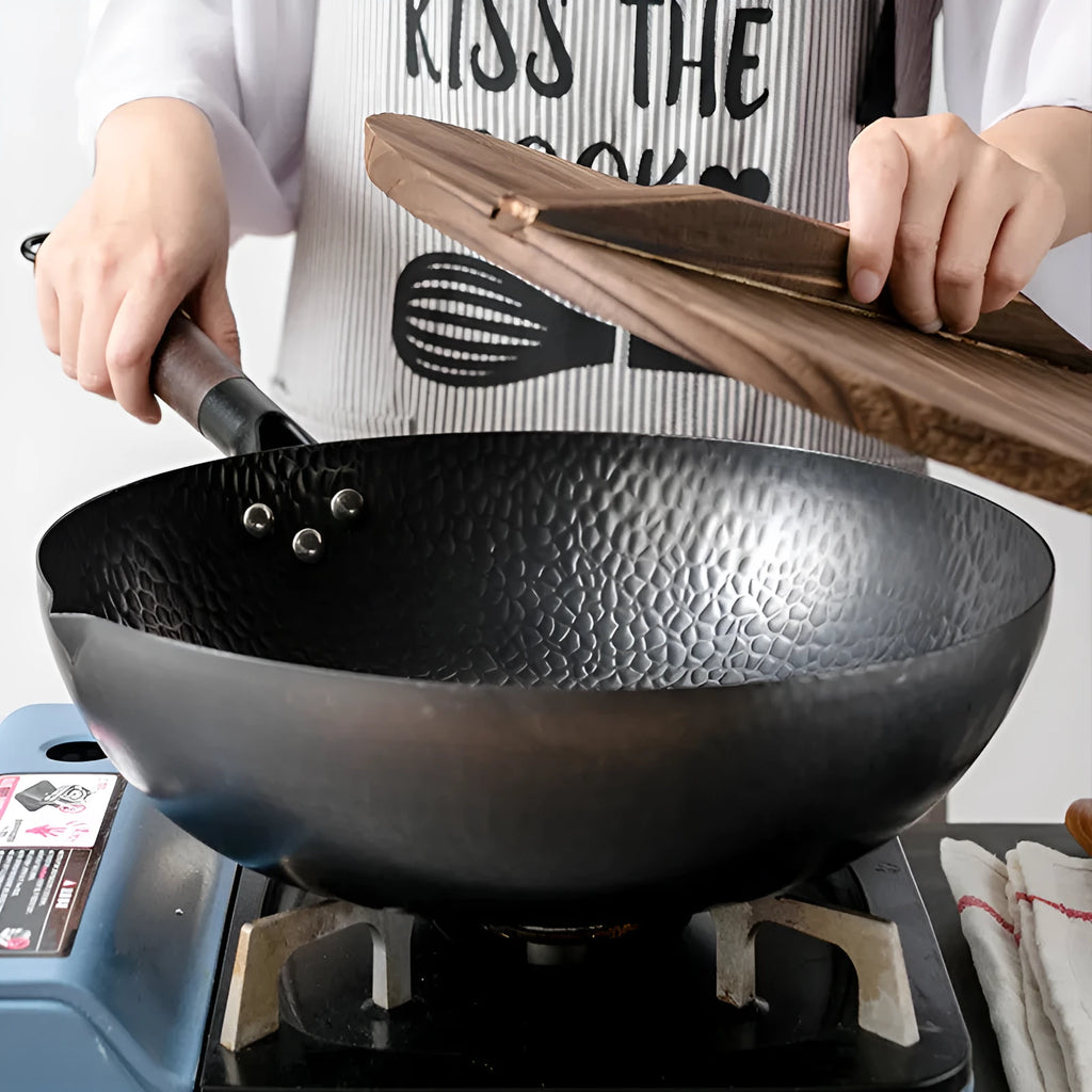 Traditional black cast iron wok with a long wooden handle, shown with a glass lid.