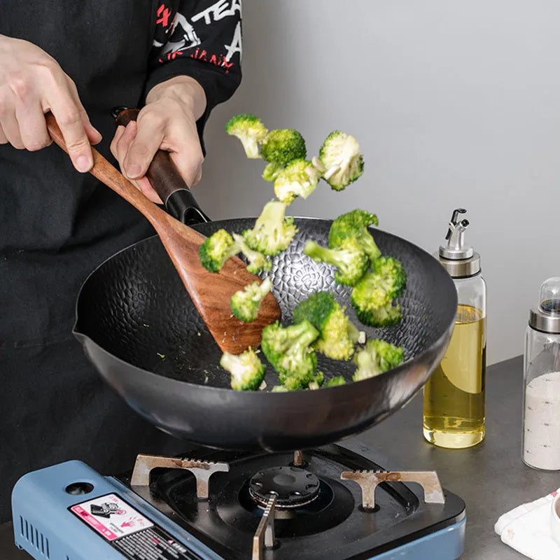 Traditional black cast iron wok with a long wooden handle, shown with a glass lid.