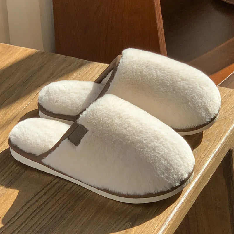A pair of solid blue plush slip-on house slippers with a non-slip PVC sole and decorative car stitching on the upper.