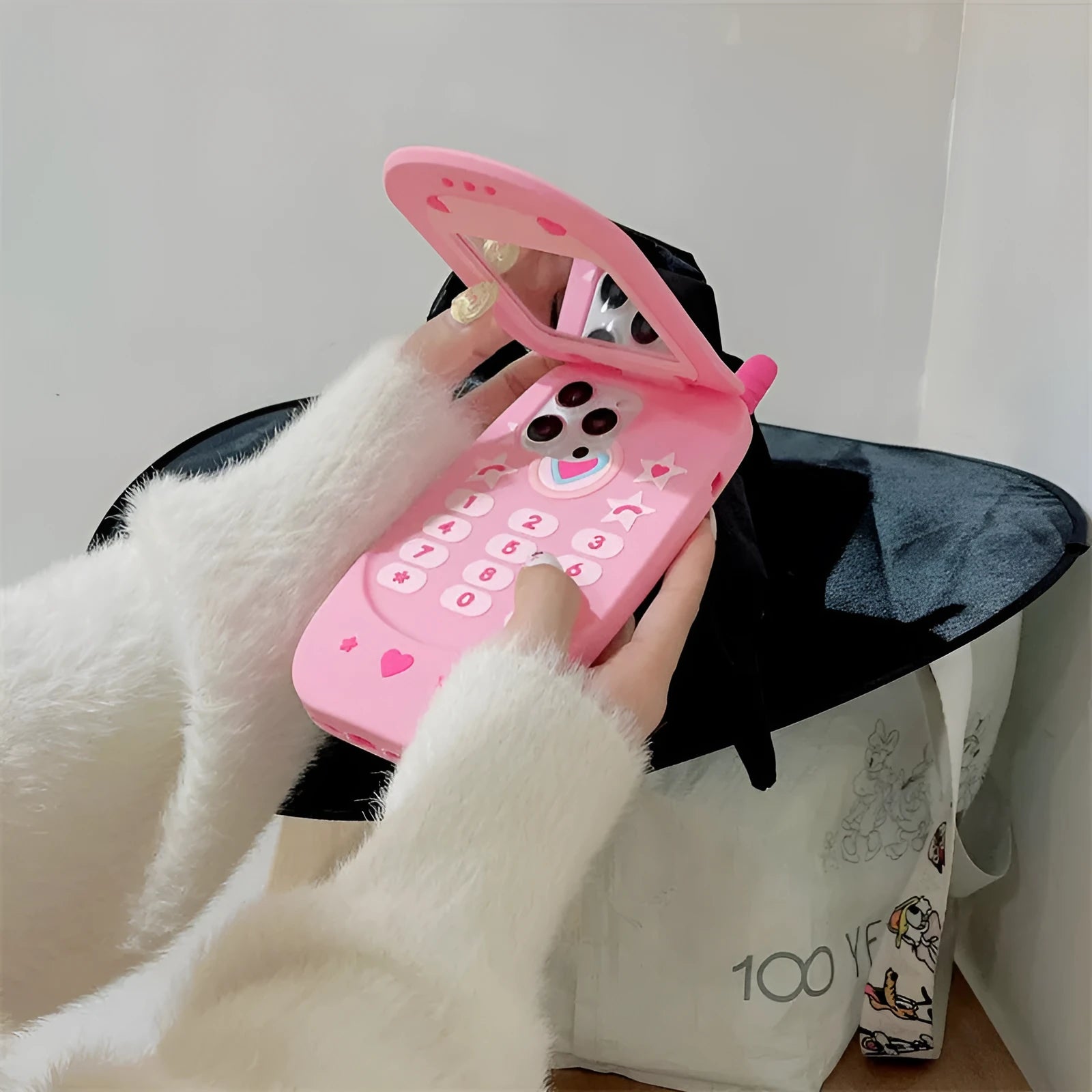 Light pink silicone flip phone case with a pop-out mirror and a cartoon charm, shown next to an iPhone.