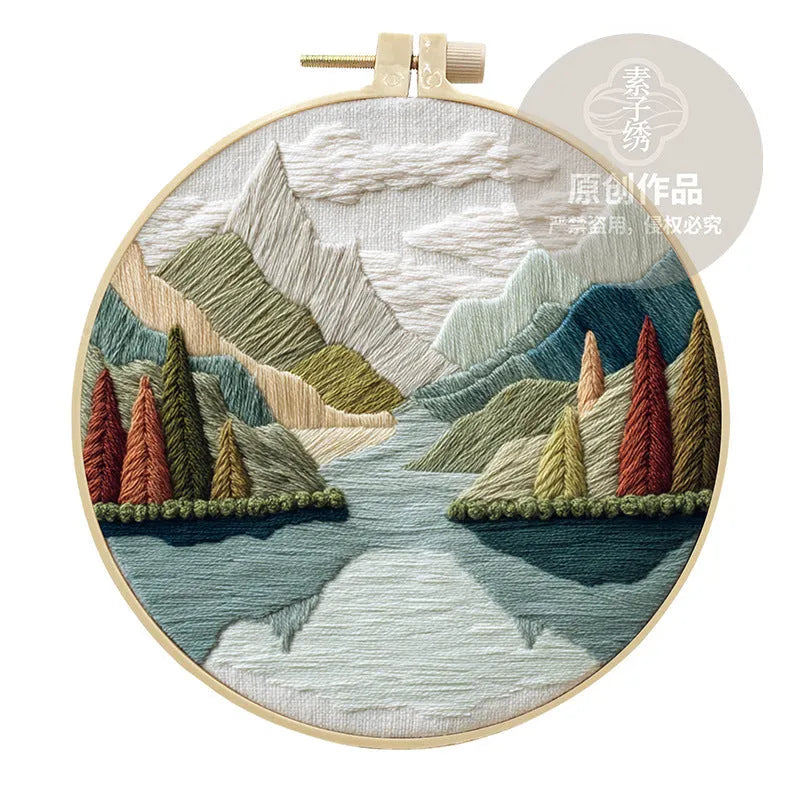 Embroidery DIY kit components: printed landscape fabric, embroidery hoop, coloured threads, and needles laid flat.