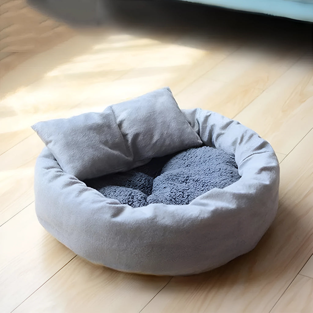 Grey round plush pet bed with raised edge and pillow, shown on a non-slip base.