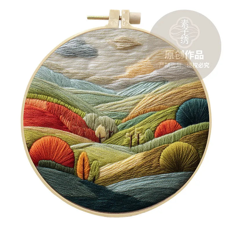 Embroidery DIY kit components: printed landscape fabric, embroidery hoop, coloured threads, and needles laid flat.